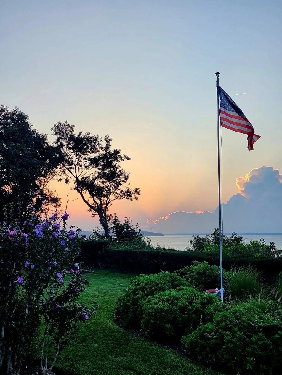 The yard at Cottage 1 at dusk, with a flag flying above flowering shrubs and a grassy path toward the bay