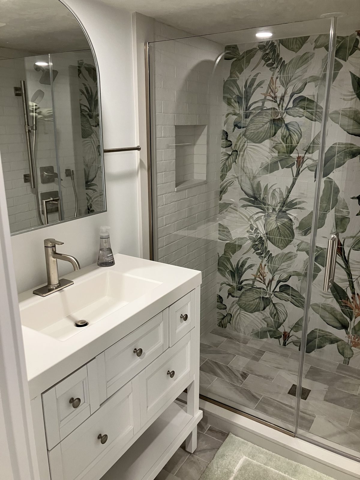 The downstairs hallway bathroom, with a walk-in shower in botanical tile, a white vanity, and an arched mirror