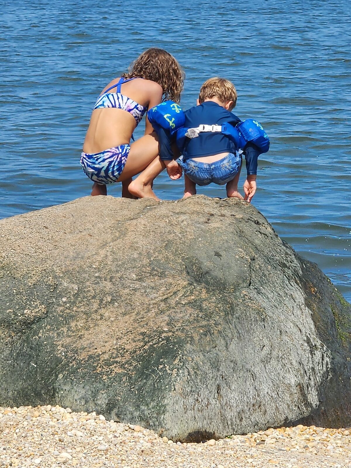 Two children crouched on a large rock at the water's edge, looking into the bay